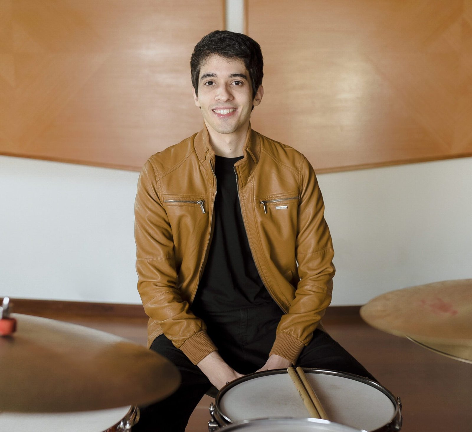 Professor Zeca Vieira vestindo jaqueta de couro marrom, sentado à bateria com as baquetas apoiadas sobre a caixa, aguardando aluno para aula de bateria.