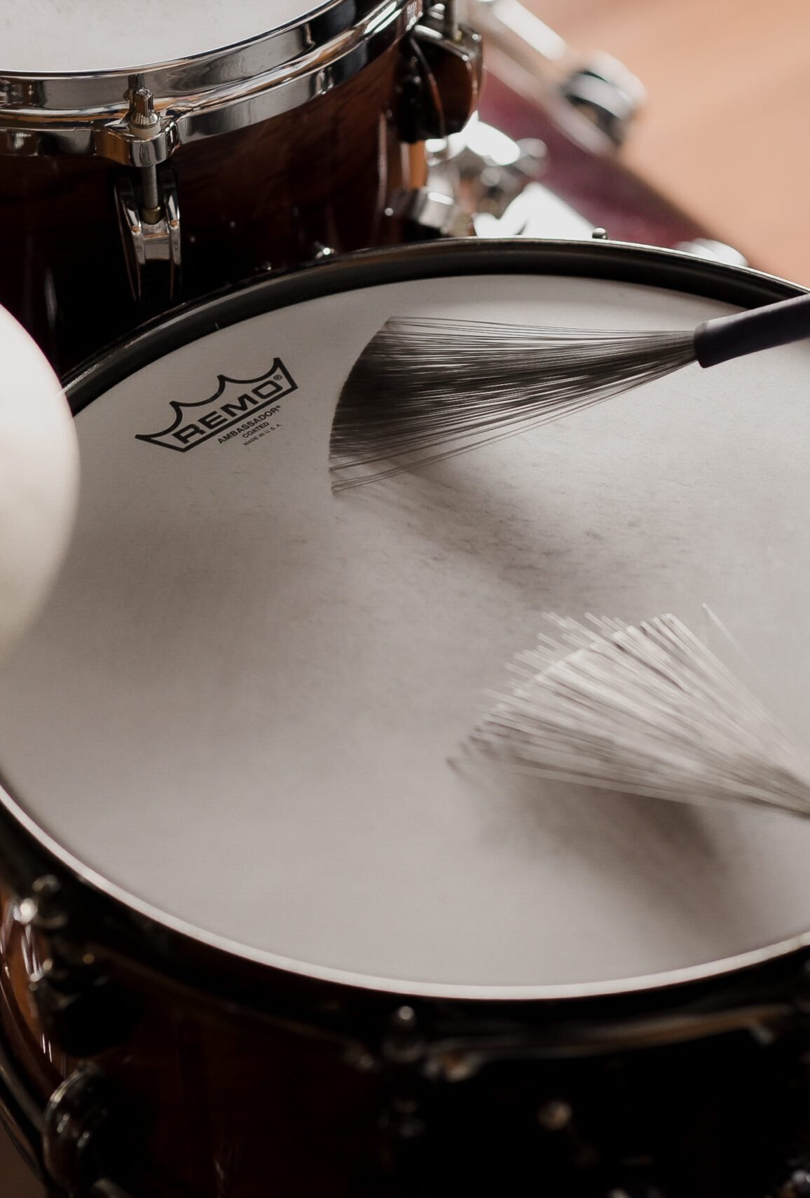 Close-up de vassourinhas (brushes) tocando a pele porosa de uma caixa Remo durante aula de bateria focada em Jazz e dinâmica suave.