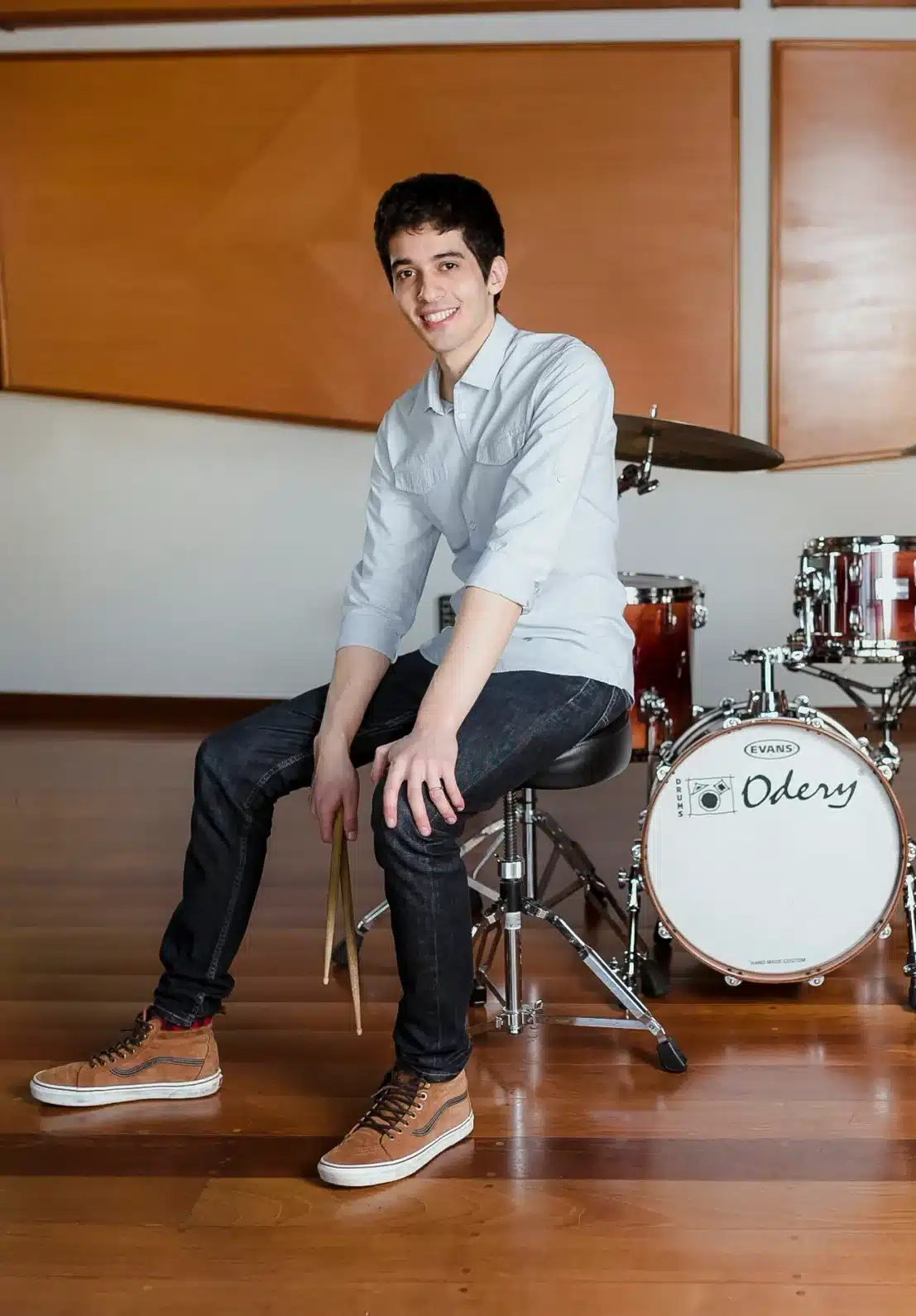Professor Zeca Vieira sentado no banco da bateria segurando baquetas, sorrindo, em um estúdio com bateria Odery preparado para aula de bateria.
