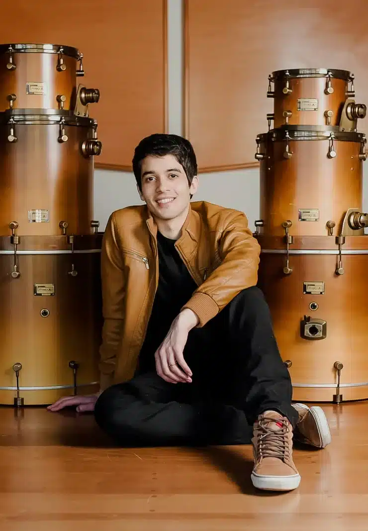 Professor Zeca Vieira sorrindo, sentado no chão do estúdio entre duas pilhas de tambores acústicos de madeira de alta qualidade, utilizados na aula de bateria.