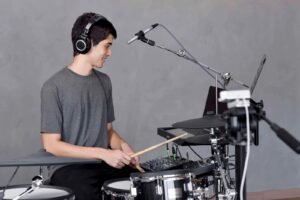 Jovem baterista sorrindo com fones de ouvido, tocando bateria eletrônica em um estúdio com microfones e câmera para transmissão de uma aula de bateria online.