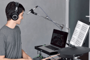 Aluno sorrindo com fones de ouvido durante uma aula de bateria campinas no formato online, acompanhando a partitura e o professor pela tela do notebook.