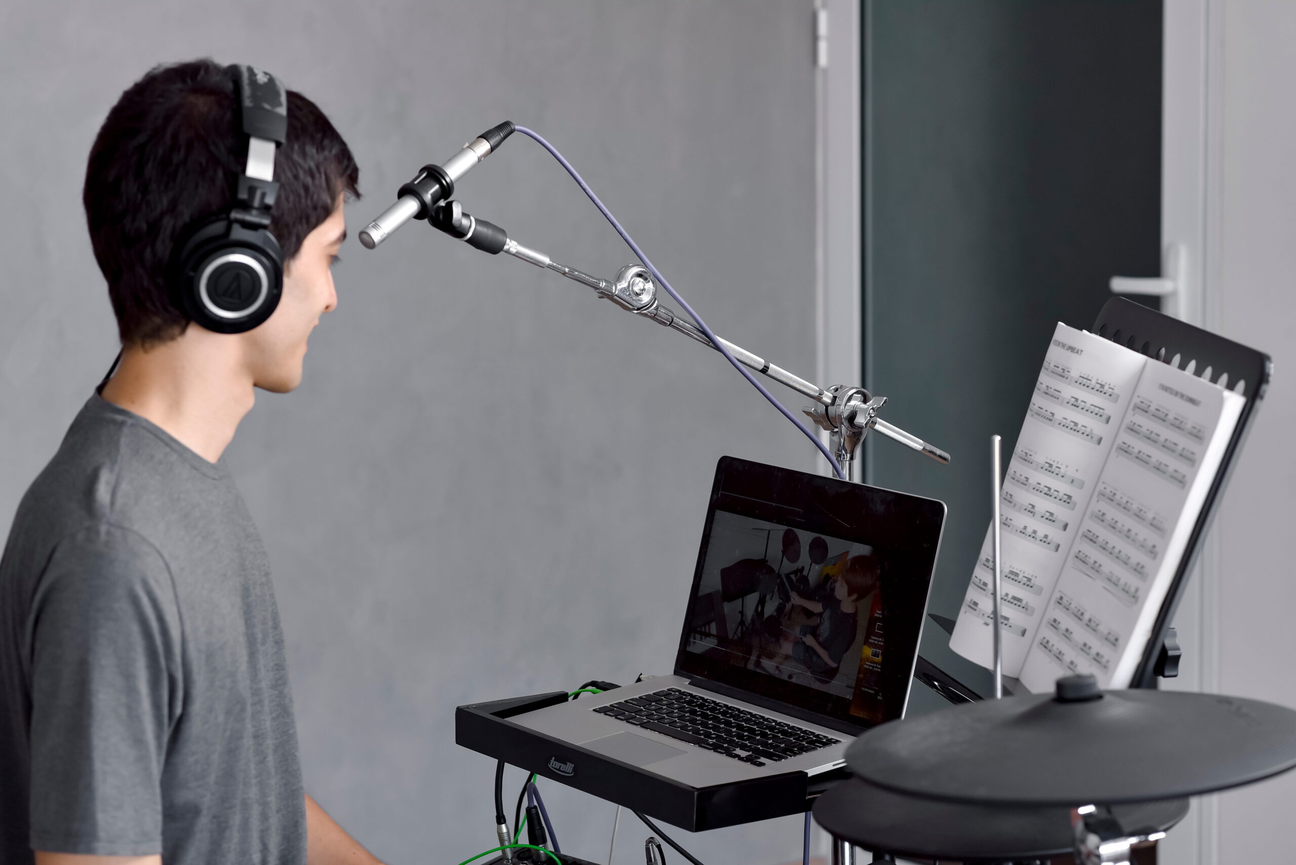 Um jovem aluno usando fones de ouvido Audio-Technica e olhando para um notebook que exibe uma aula de bateria online, posicionado atrás de uma bateria eletrônica com estante de partituras visível